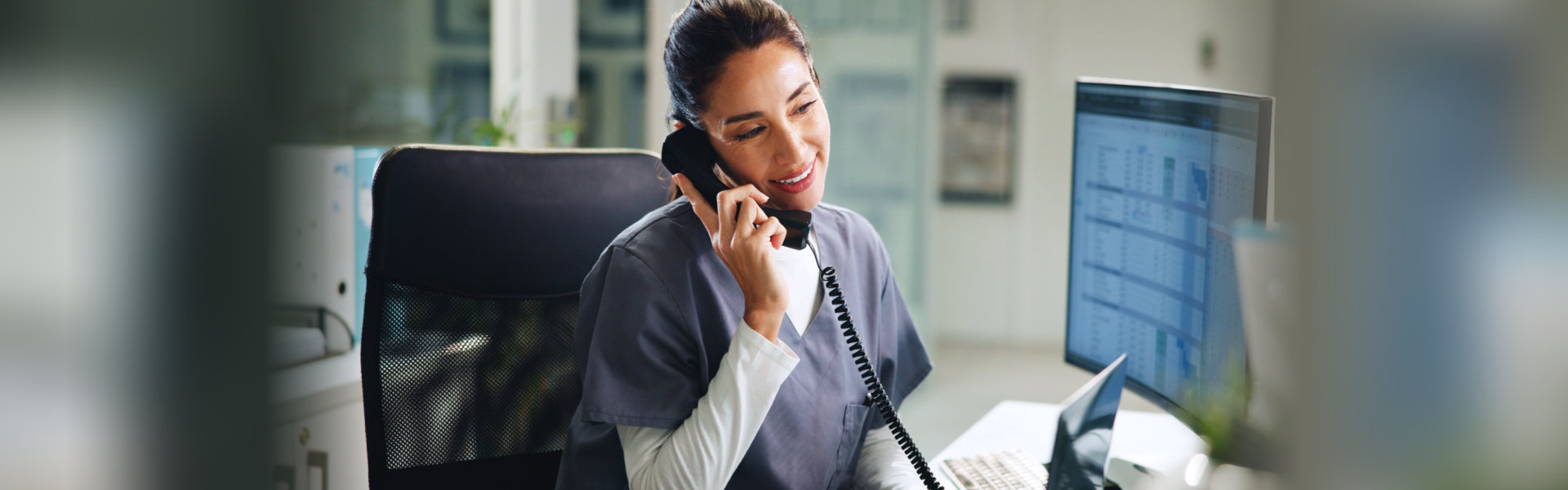 Smiling healthcare staff member in scrubs answering a phone call at her desk
