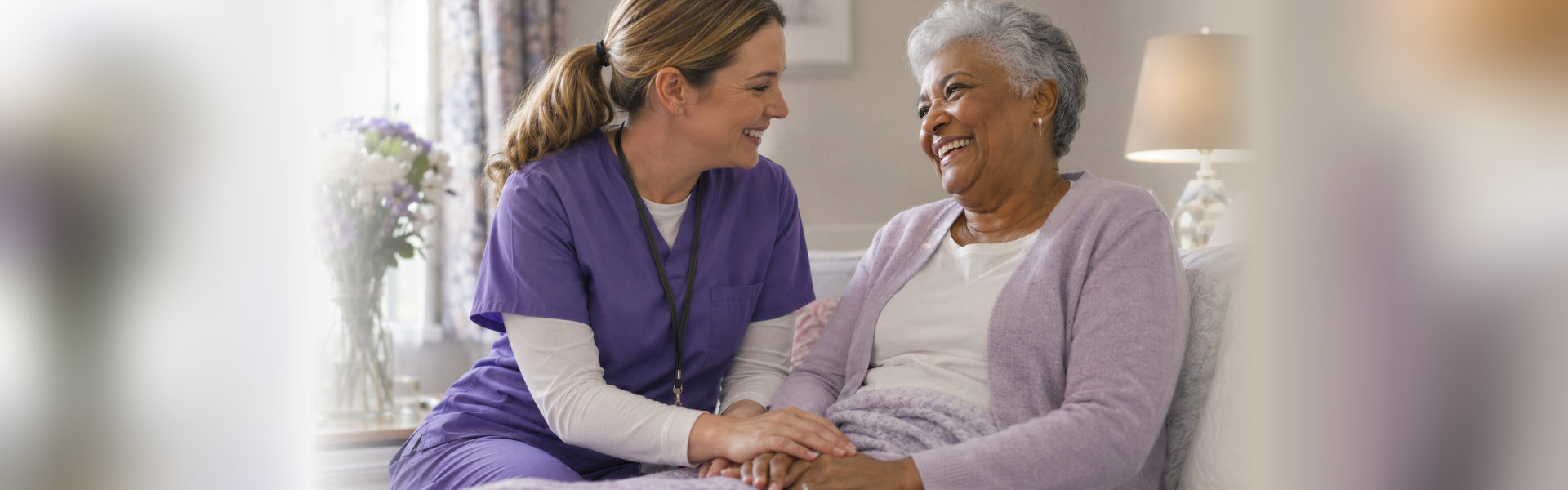 Smiling caregiver in purple scrubs holding the hands of a laughing elderly woman